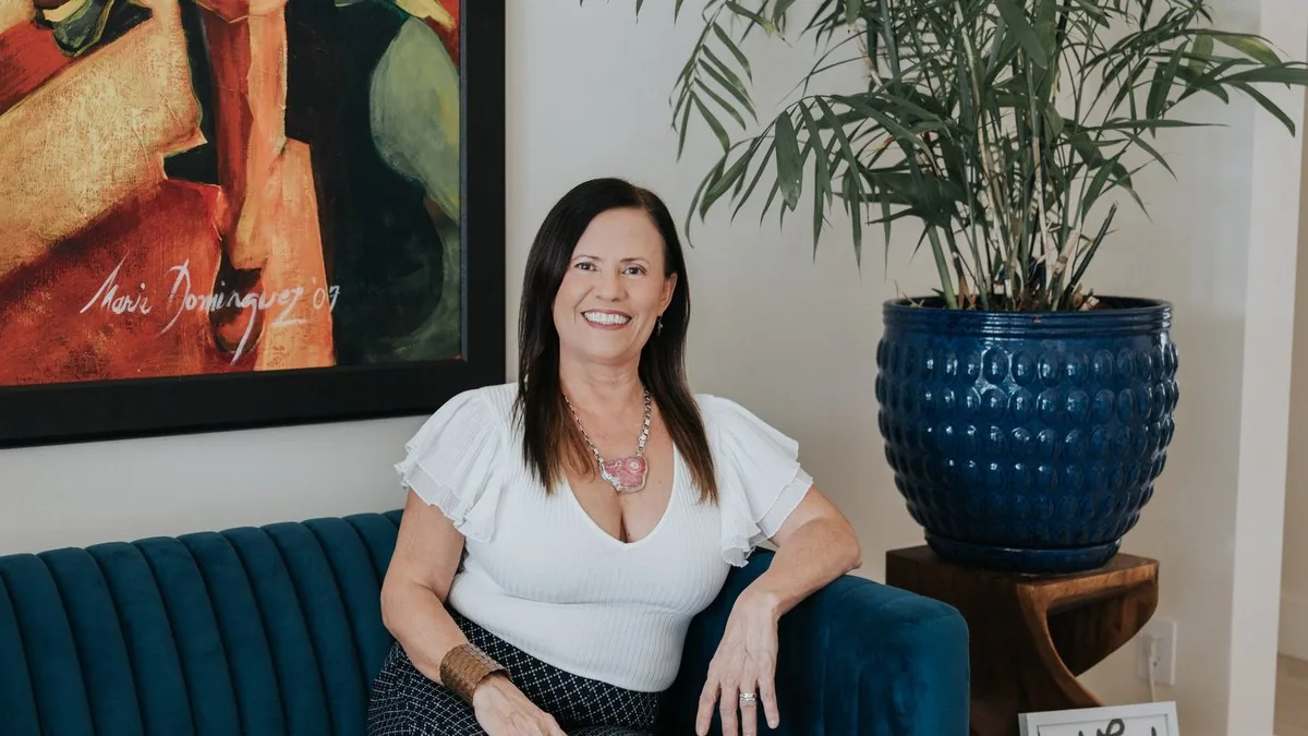 Isa Herrera seated on a deep teal sofa in a white top, beside a framed painting and a tall palm.