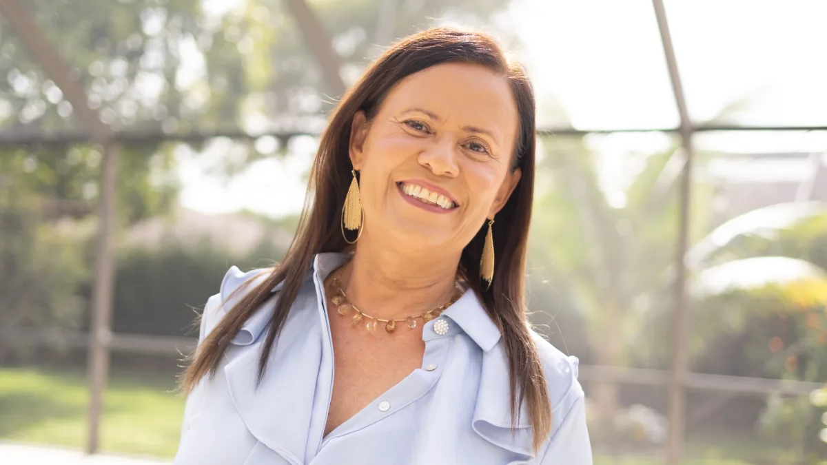 Isa Herrera, MSPT, CSCS, smiling in a pale blue shirt outdoors, backlit by sun through a screened garden.